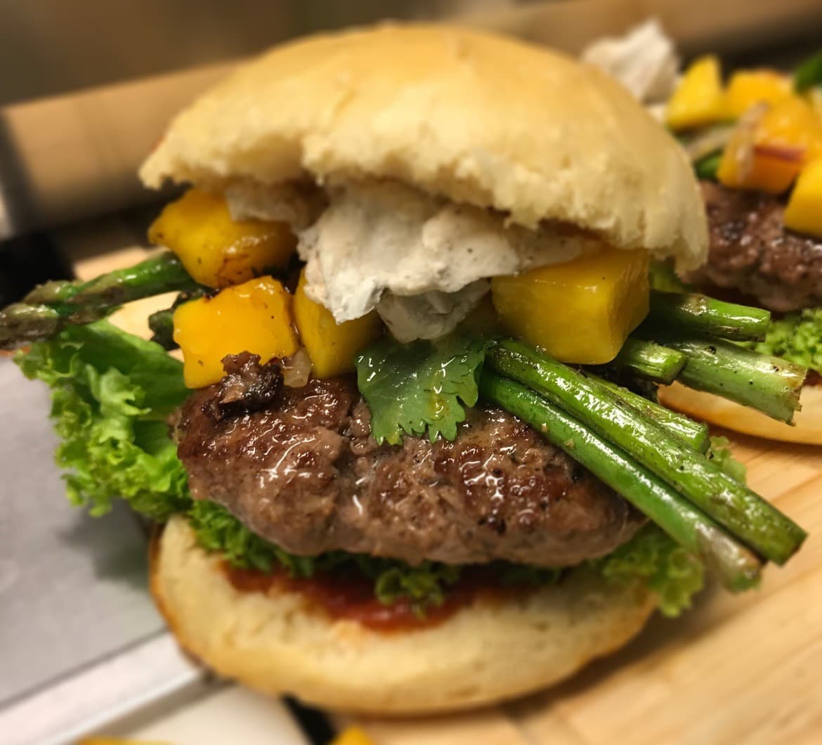 Nahaufnahme eines Burgers: Patty mit gelben Fruchtwürfeln, grünen Gemüsestangen, Kräutern und weißer Creme im Bun