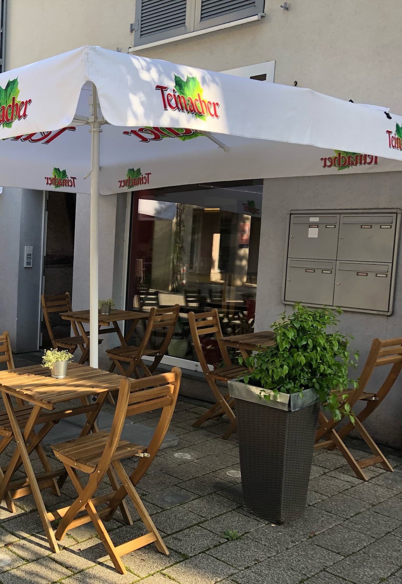 Terrasse vor dem Lokal mit Holztischen, Klappstühlen und einem Pflanzkübel